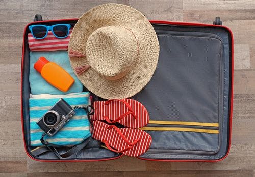 A suitcase open on the floor with sunglasses, a straw hat, a camera, flip flops, sunscreen, and beach towels