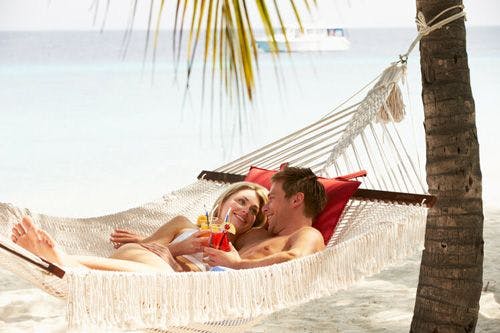 A couple lying in a hammock by the sea drinking cocktails