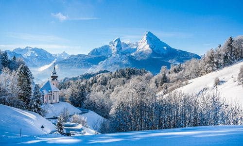 A winter scene in Europe with snow-covered fields and mountains and a church on a hill