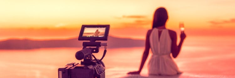 A woman sitting on a wall with a glass of champagne watching the sunset, while a movie camera films her
