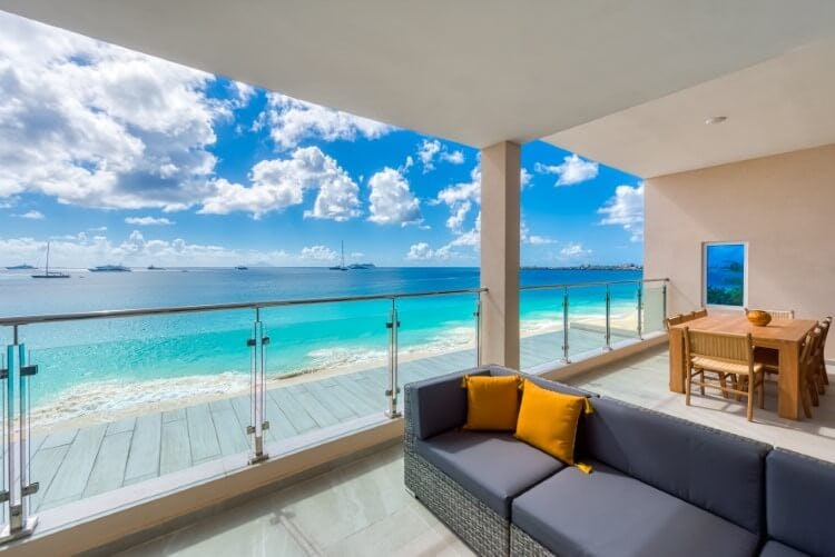 C'Scape penthouse rental - image of a balcony area with a comfortable grey couch with mustard-yellow cushions, a table and chair set, and a glass barrier overlooking a white sand beach and beautiful blue water