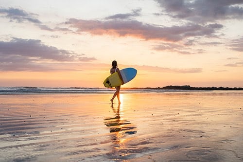 A woman carrying and blue and yellow surfboard along the beach at sunset
