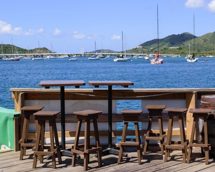 A waterfront restaurant in the Caribbean, with wooden tables and chairs overlooking sailboats in the harbor
