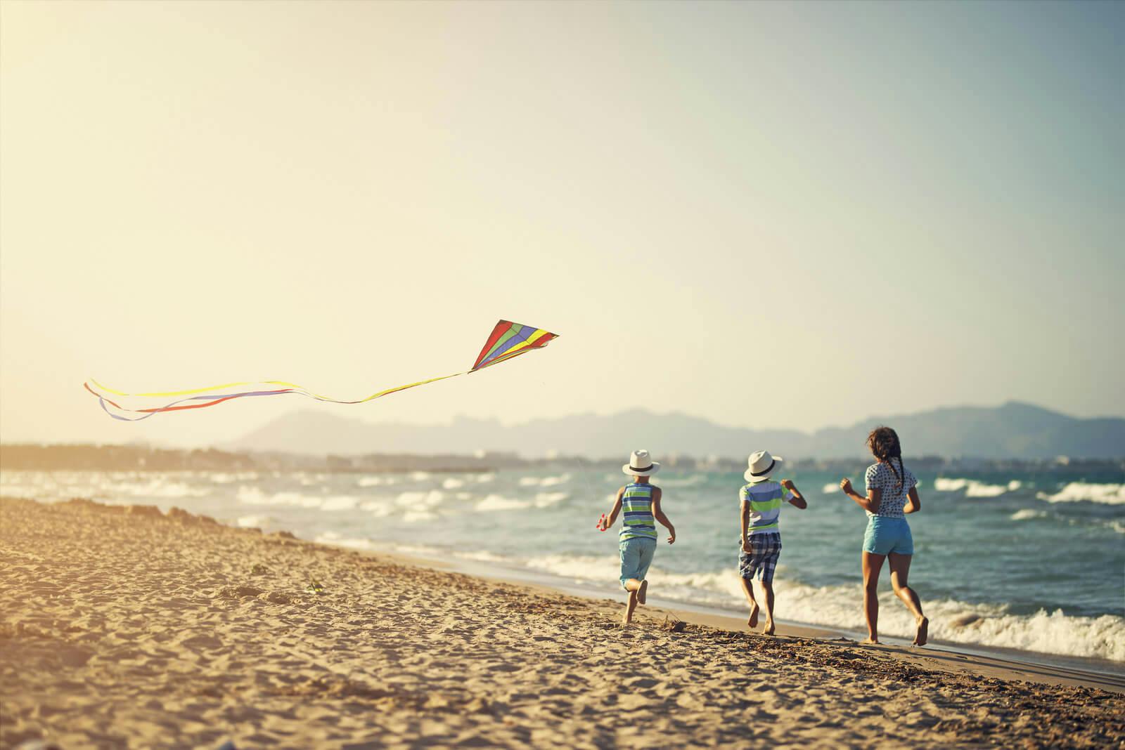 Three children run along a white sand beach flying a kite