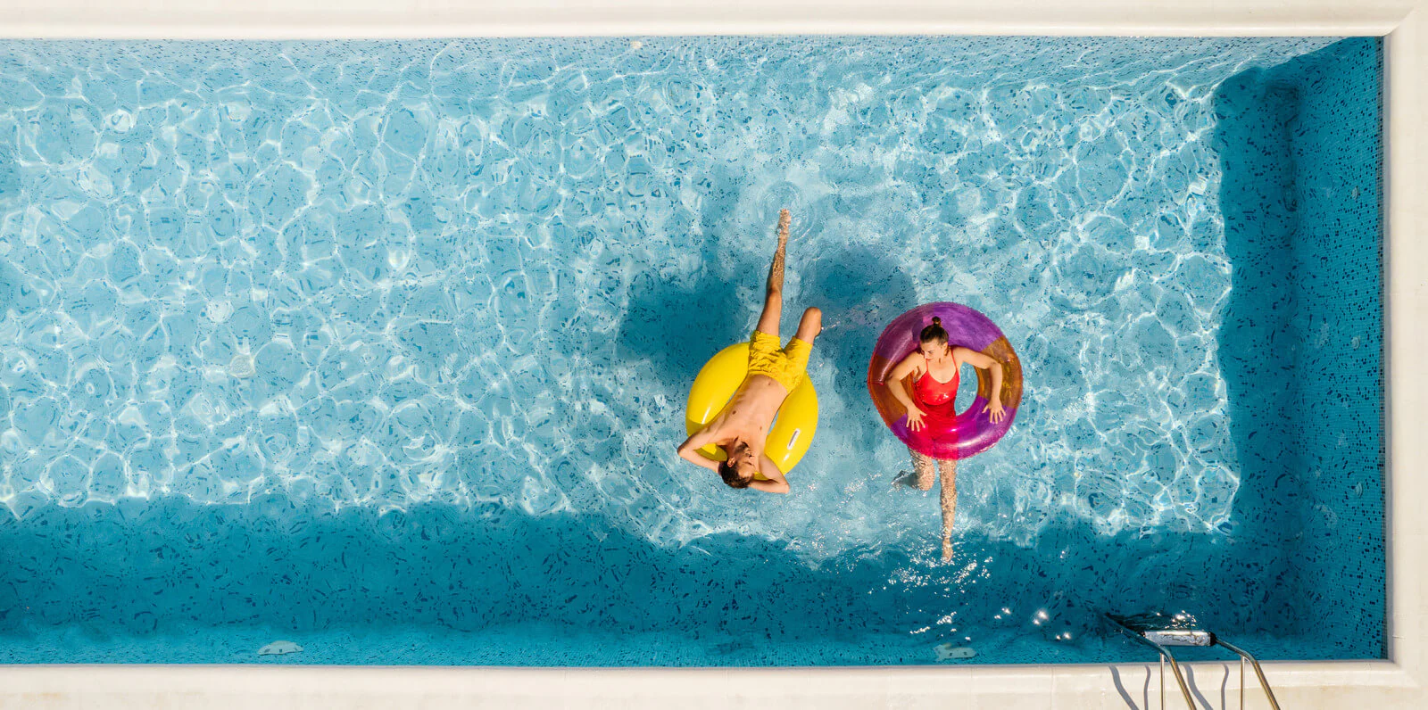 Two people, a man and a woman, floating on inflatable rings in a pool. The man wears yellow trunks on a yellow ring, while the woman wears a red bathing suit on a red ring
