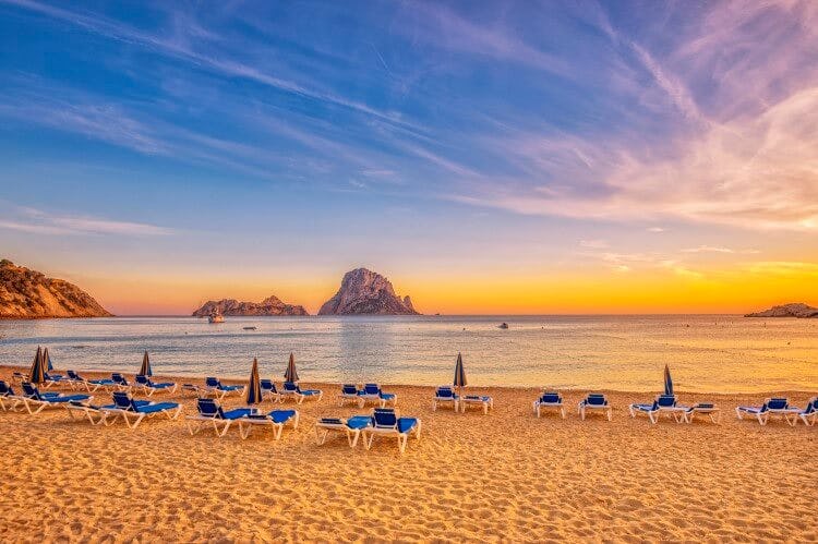 A white sand beach in Ibiza with rock formations in the water and sun loungers on the sand