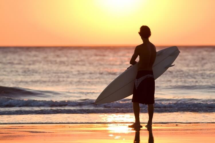 Surfing in the Caribbean - a young man with a surfboard silhouetted against the setting sun in front of the sea