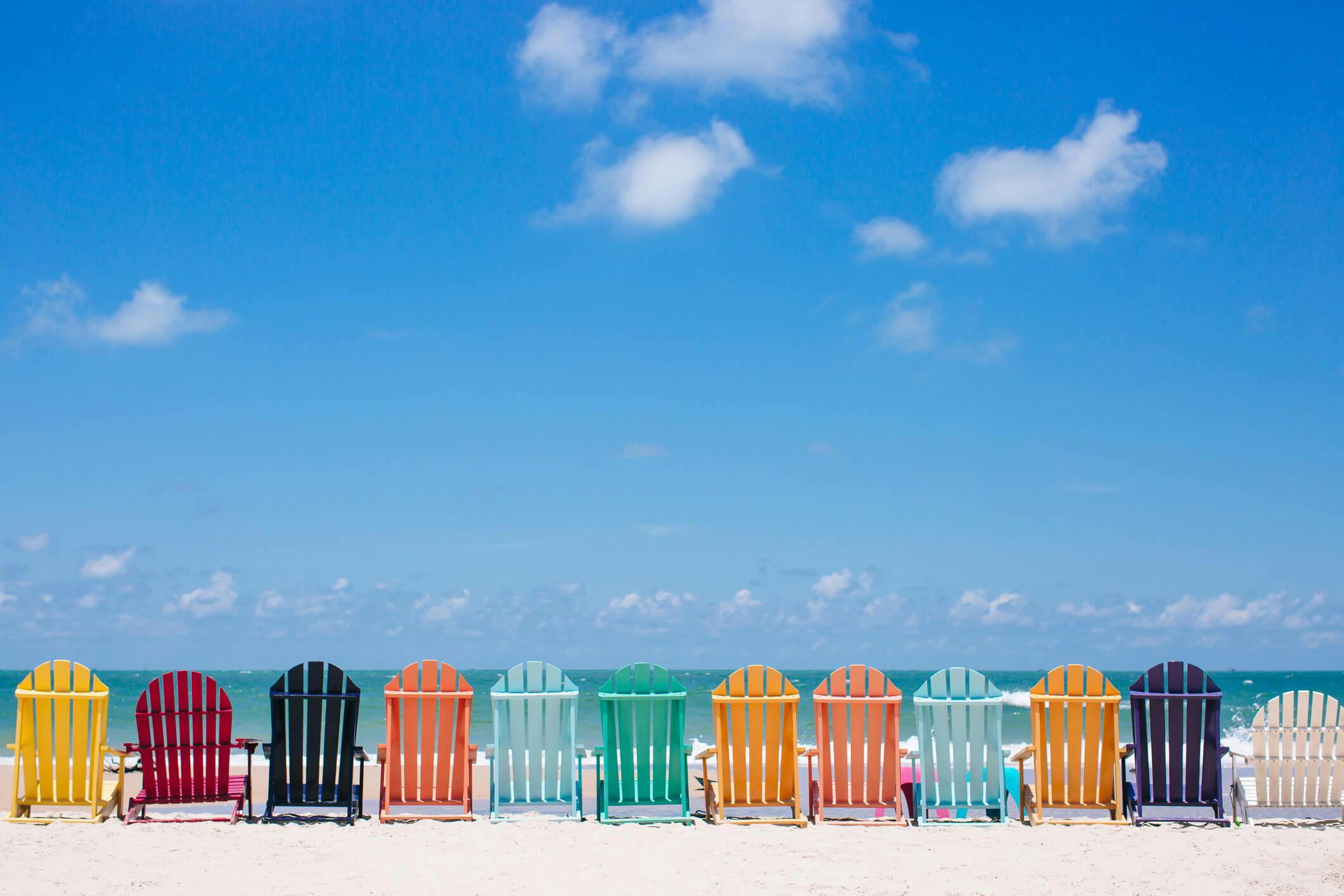 Colorful wooden beach chairs arranges in a line along a white sand beach facing the sea