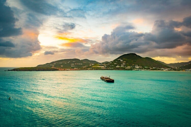 Sea view of Saint Martin with a small ship sailing towards green forested hills over bright blue water, with the soft colors of a sunset blazing in the late afternoon sky
