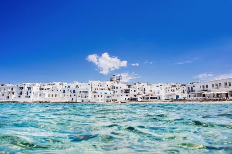 Naoussa village in Paros, Greece, with white sugar cube buildings along the harbor and clear blue sea in front 