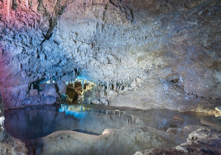 The inside of Harrison Cave in Barbados, with stalactites and a still pool of water in the center. The walls of the cave are lit with soft blue and pink lighting