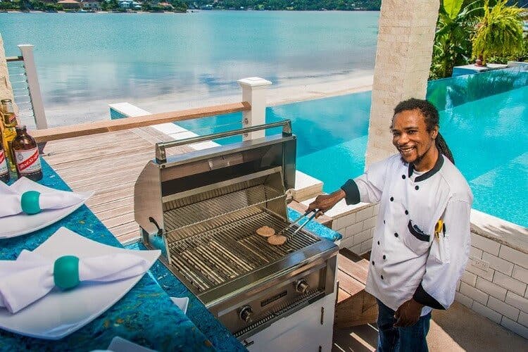 A young male member of staff cooking burgers on a barbeque in front of a swimming pool in a Caribbean vacation home