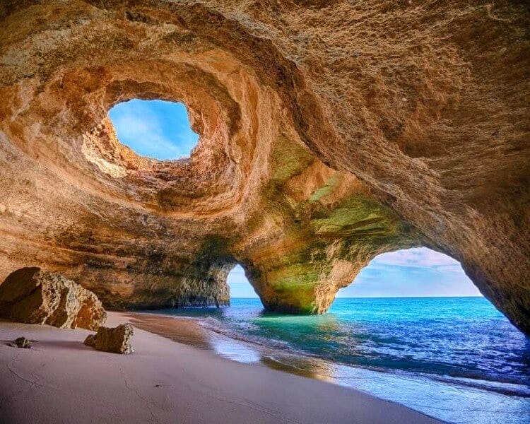 Benagil Cave in the Algarve, an arched rocky cave with a round hole in the ceiling and sea water flowing onto a white sandy beach inside