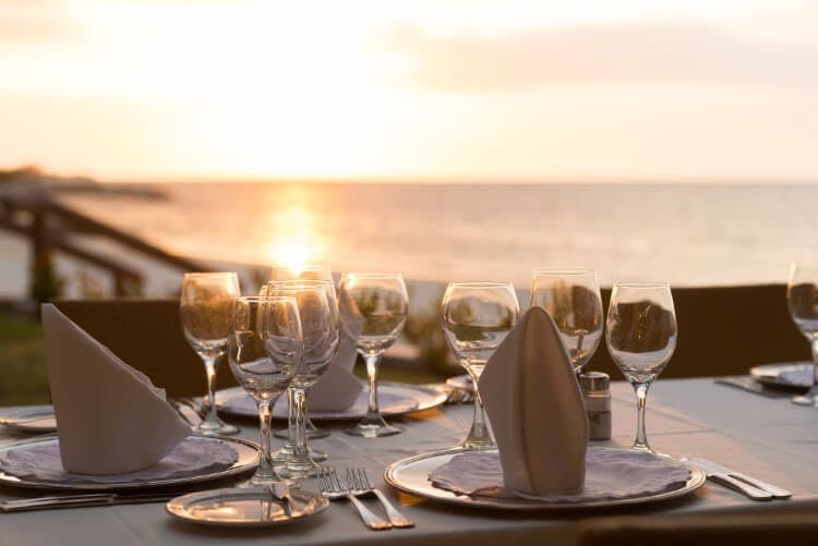 A table set for a romantic dinner in a beachfront Caribbean restaurant, with the sun setting over the sea in the background