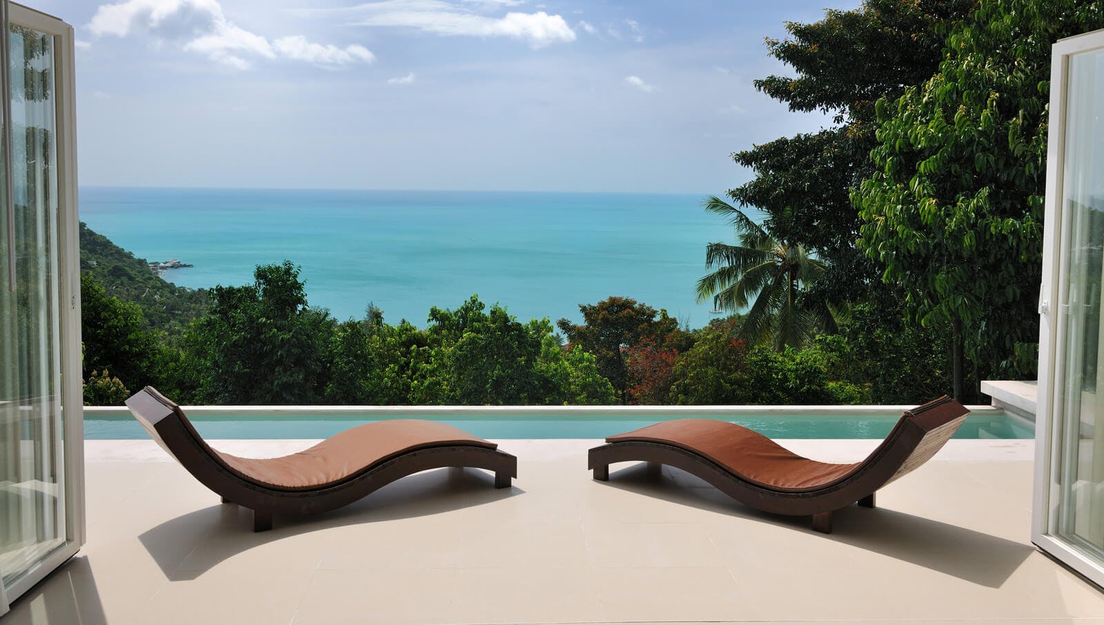 two brown sun lungers on a patio by a private pool overlooking trees and the sea