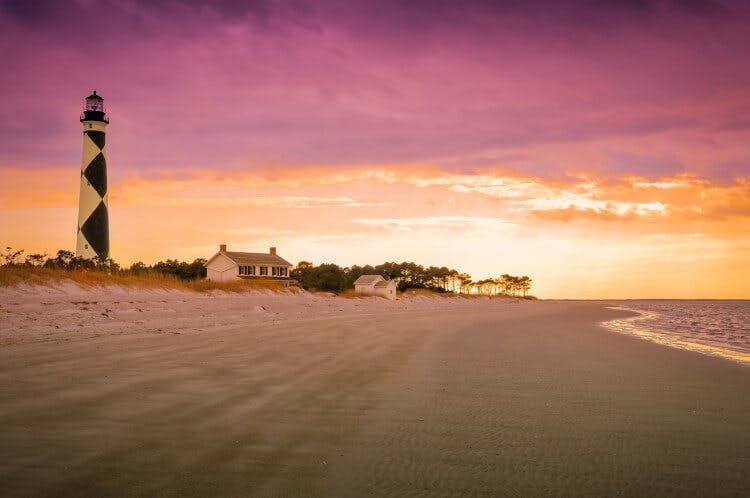A purple and orange sunset over a white sand beach with a small white building and a black and white lighthouse