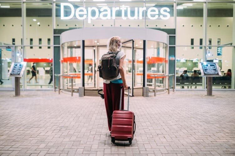 A young woman pulls a red wheelie suitcase behind her as she enter an airport departures hall
