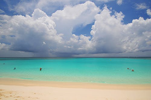 A white sand beach in Anguilla with people swimming in the sea and fluffy white clouds in the sky