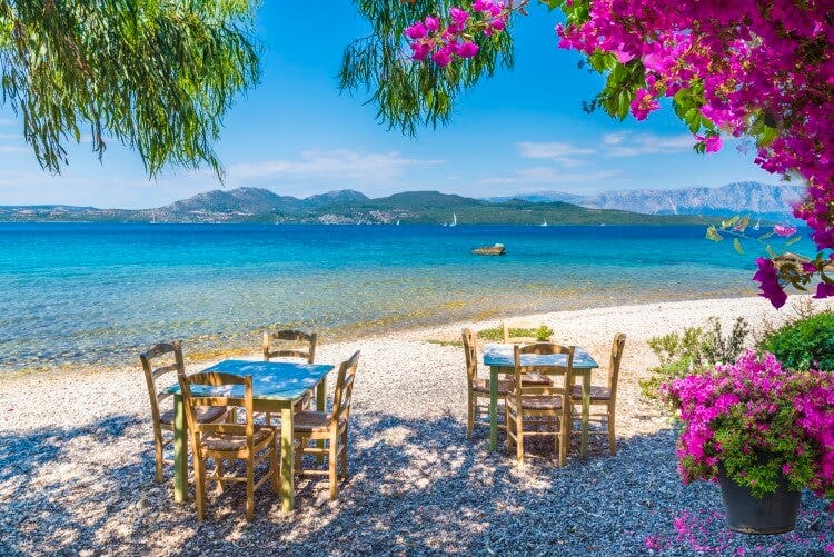 A Greek taverna with tables and chairs set out on a white sand beach with pink bougainvillea 
