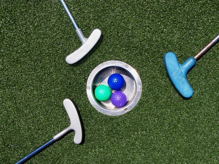 Three mini golf clubs encircle a hole in which three colorful golf balls reside