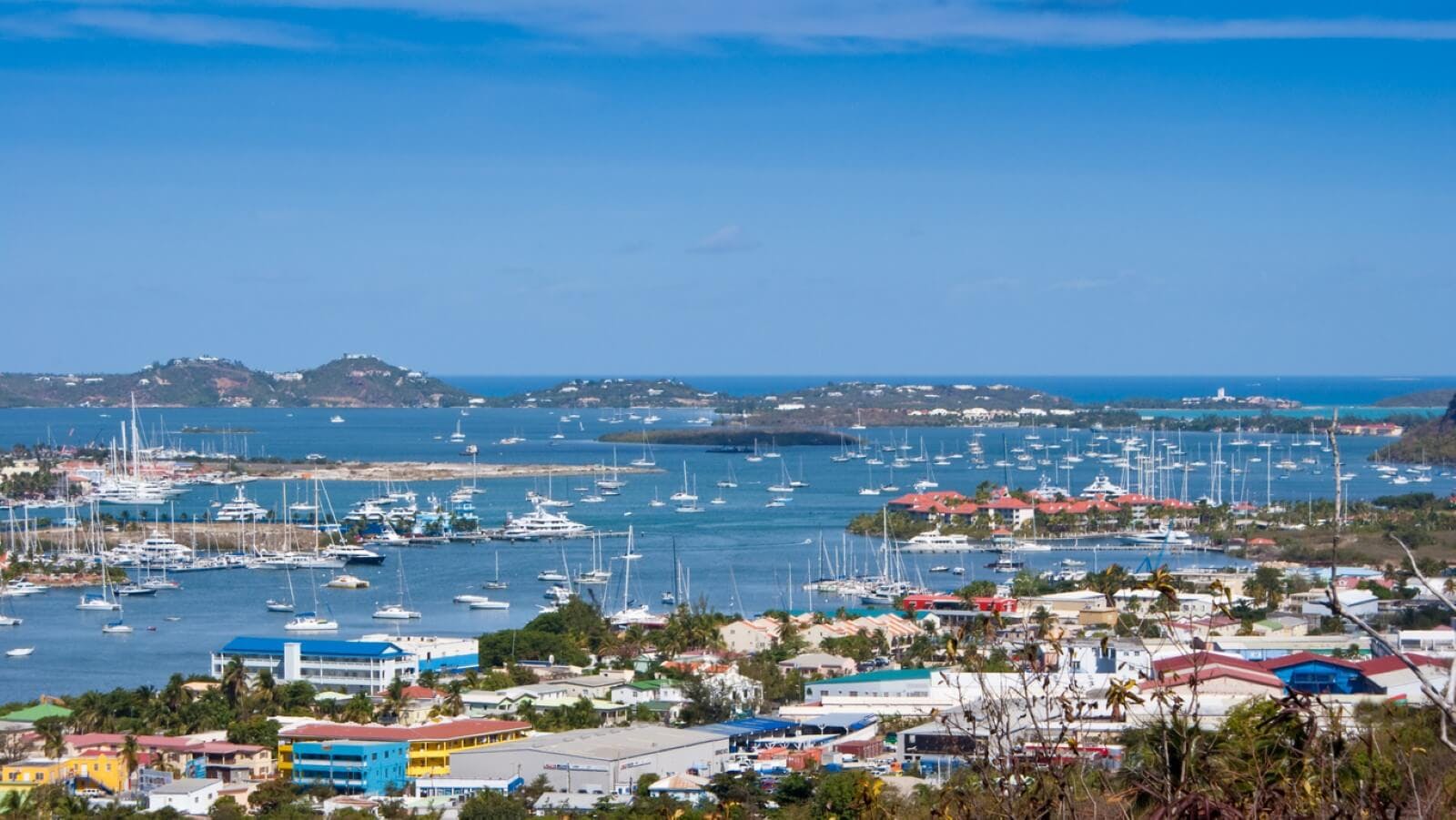 Simpson Bay harbor in Saint Martin, with fishing and leisure boats and yachts in clear blue sea, green hills in the background and small colorful buildings lining the shore