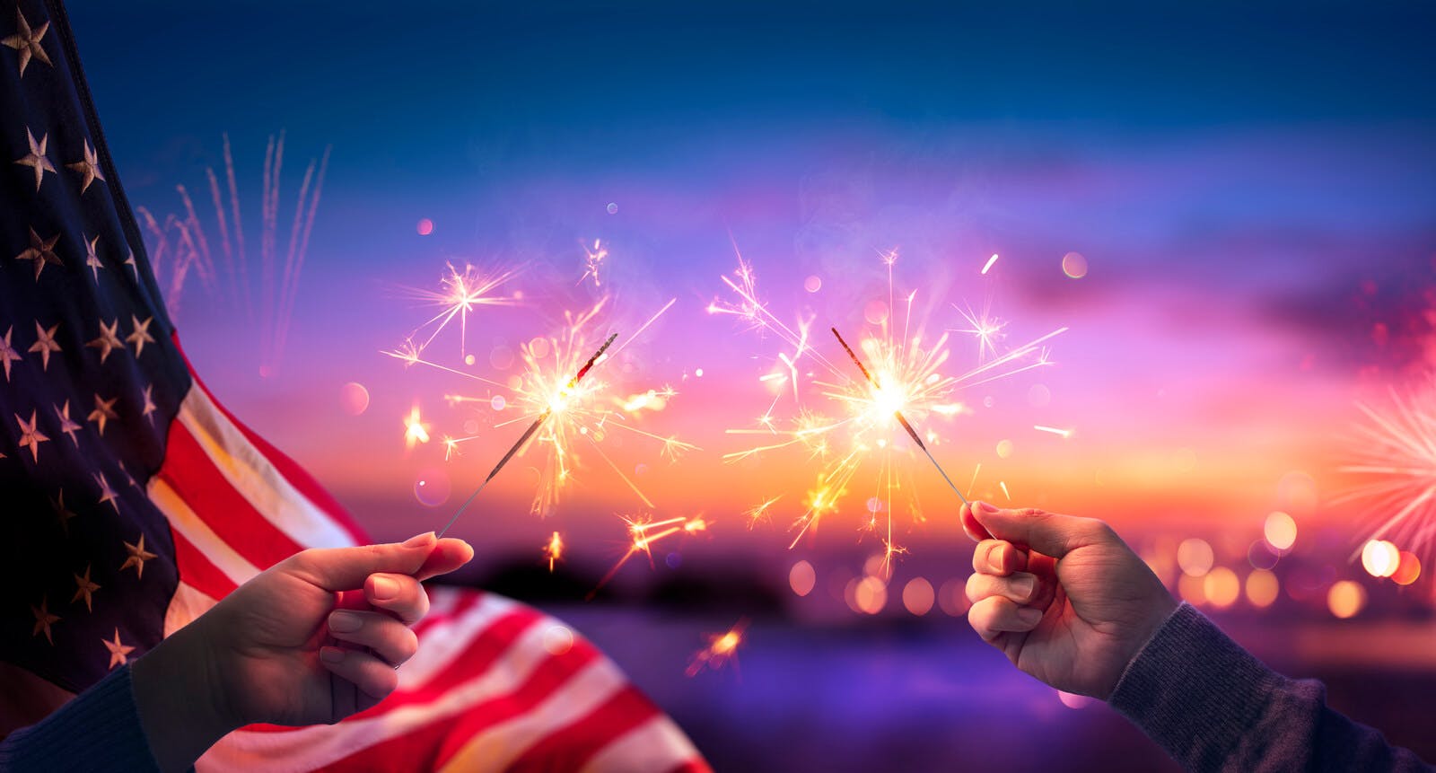 A sunset sky with two children's hands holding sparklers in front of an American flag