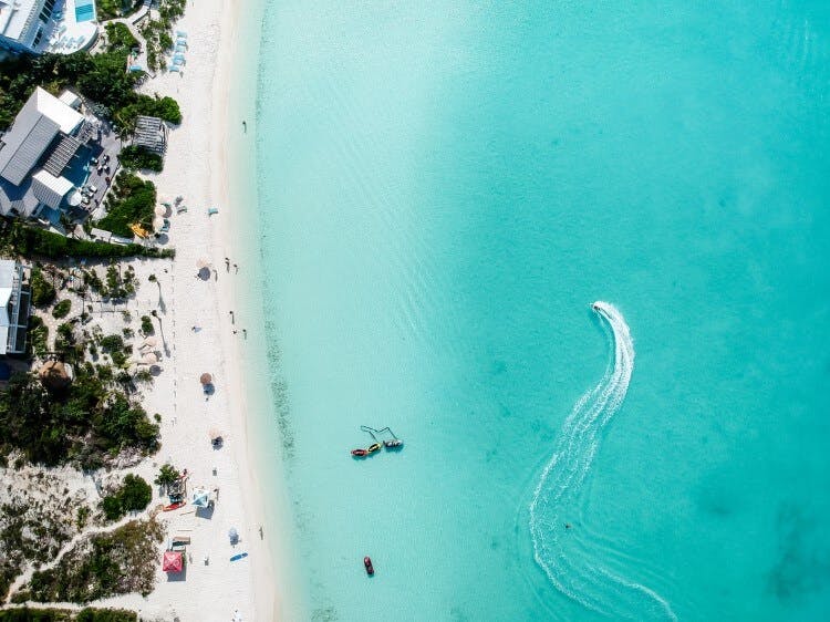 A drone shot of Sapodilla Beach, showing clean white sand, bright turquoise water, with a speed boat leaving a curved wake behind in, and vacation rentals lining the shore