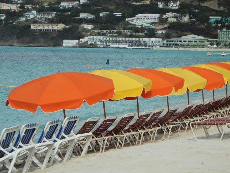 Saint Martin beach with a line of sun loungers with orange and yellow parasols on front of calm water