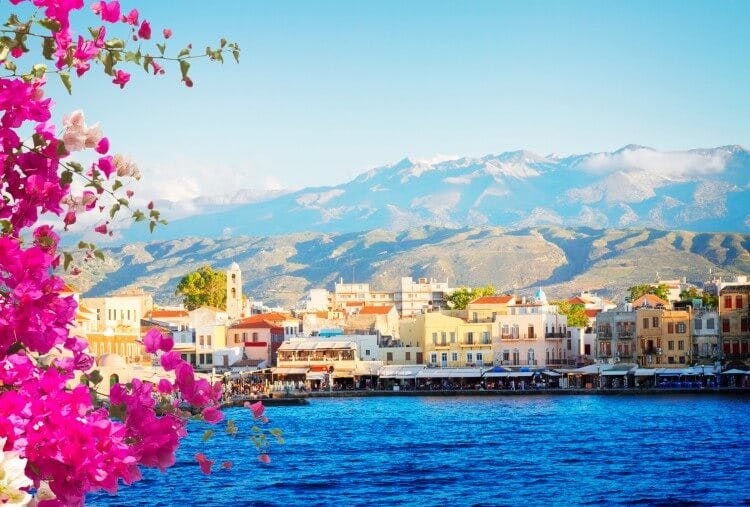 The city of Chania is Crete, with colorful cube-shaped buildings lining the waterfront and mountains in the background