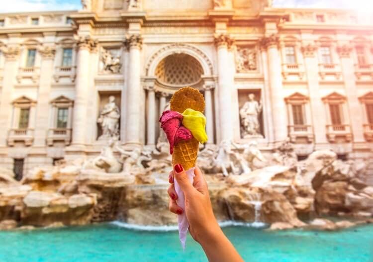 A woman's hand holding up a cone with two scoops of pink and yellow gelato in front of the Trevi Fountain in Rome