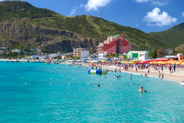 A beach in Saint Martin, with white sand, colorful buildings along the shoreline and people swimming and relaxing on the beach
