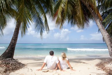 A couple sits on a white sand beach in Barbados with palm trees