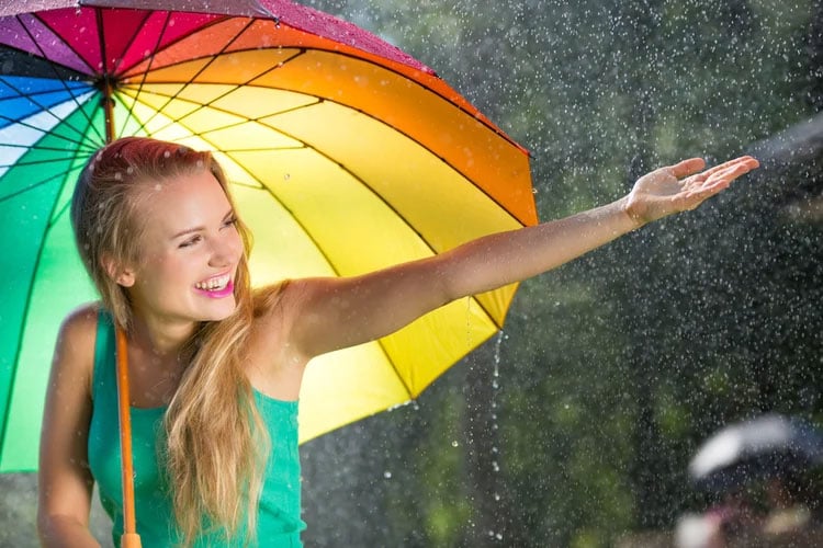 A young woman under a rainbow umbrella holding her hand out the rain