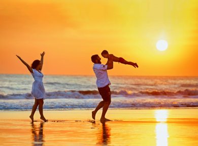 A family on a beach at sunset - a man lifts a child into the air as the woman raises her arms