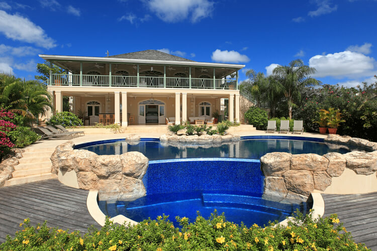 A two-story villa overlooking a large pool surrounded by palm trees and tropical plants.