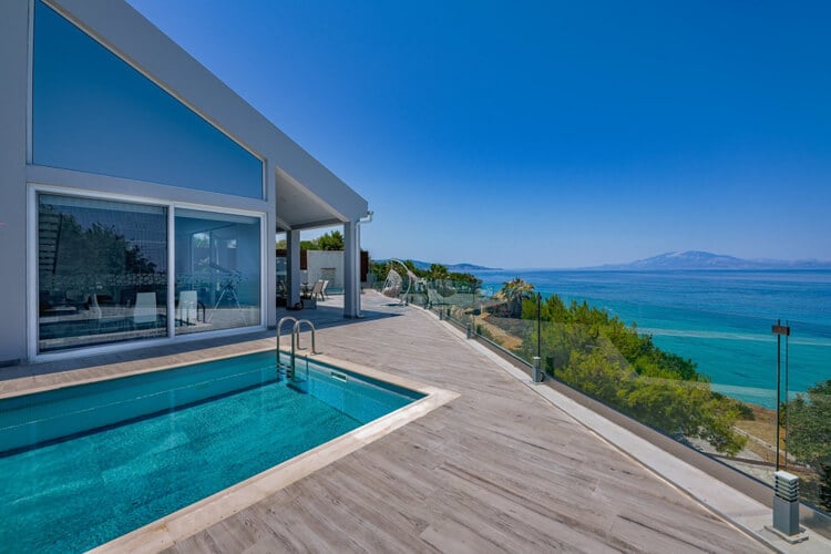 A villa, swimming pool, and wooden deck surrounded by a glass barrier with the ocean and an island in the distance 