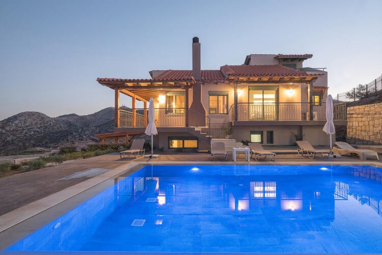 A warm villa with its lights on, mountains in the background and a pool in the foreground