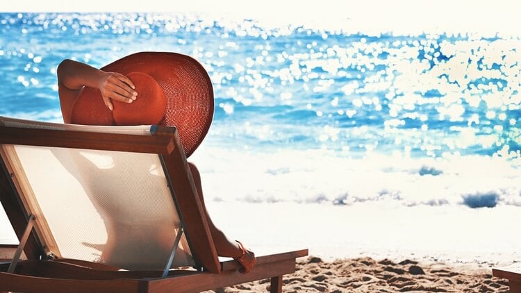 A person in a large sun hat sitting in a sun lounger on the beach looking out to sea