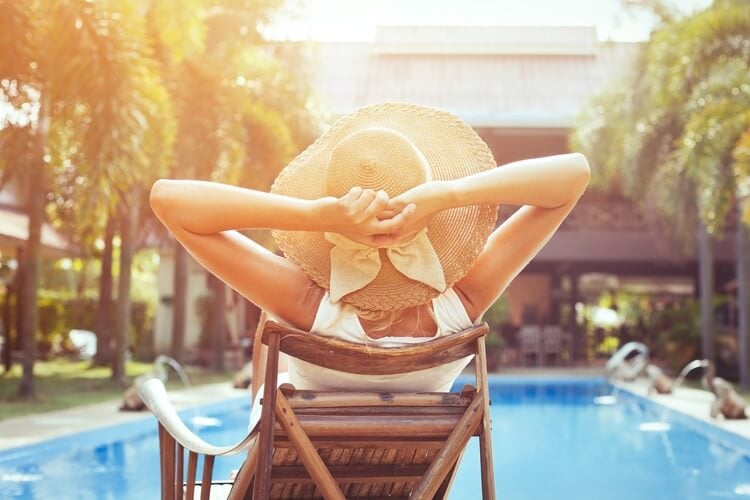 A person in a a large sun hat sits in a sun loungers facing a large pool