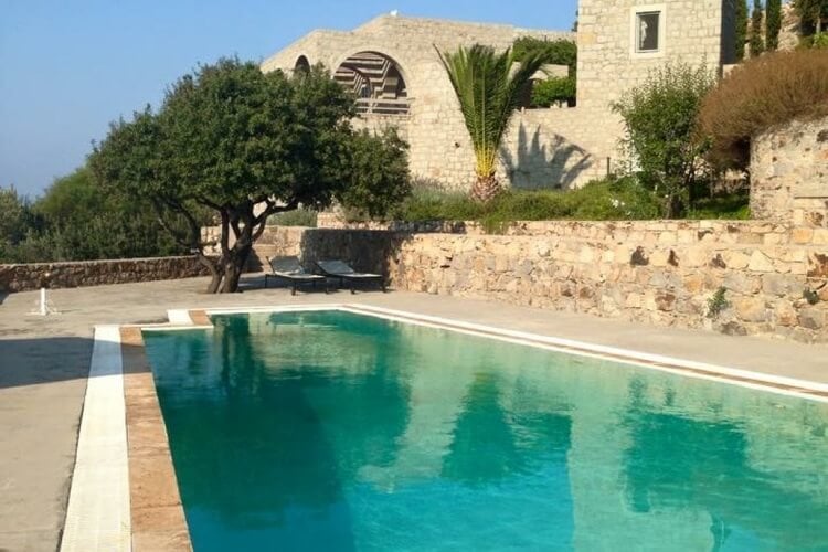 A rustic villa made out of stone, with a large pool, several trees, and sun loungers