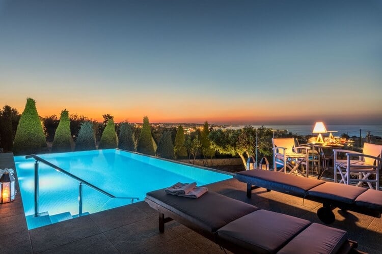 A pool, sun loungers, and dining table at dusk, with the lit up coast beyond. 