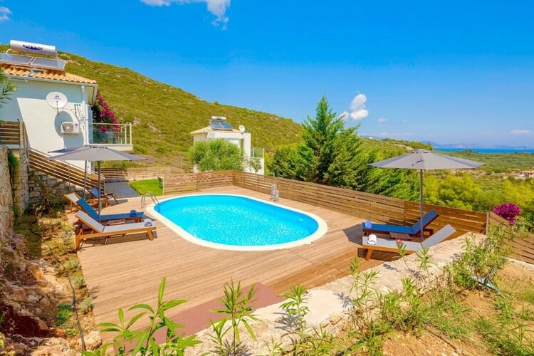 A small villa against a green hill, with a pool in the foreground and ocean in the distance.