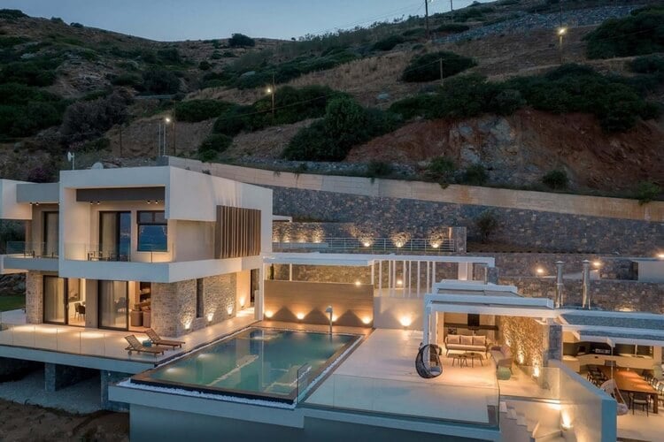 A modern villa nestled against a hill, lit up in warm orange with an infinity pool, dining area, and sun loungers.