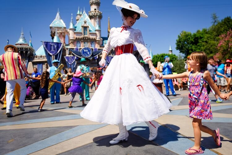 A person dressed as Mary Poppins in white dress and hat holds a child's hands as she walks in front of the Disneyland castle