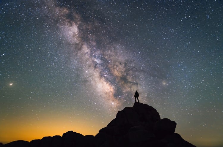 A man stands silhouetted against a clear night sky, with the Milky Way stretching overhead