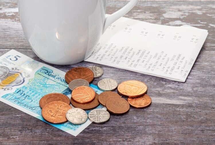 A collection of UK pennies and a five pound note left as a tip on a wooden table with a bill under a cup