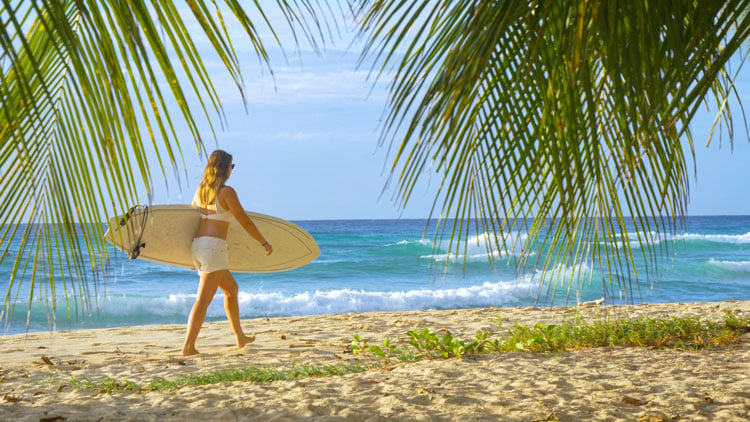 The best places to visit in December - Person carrying surfboard along golden sand beach