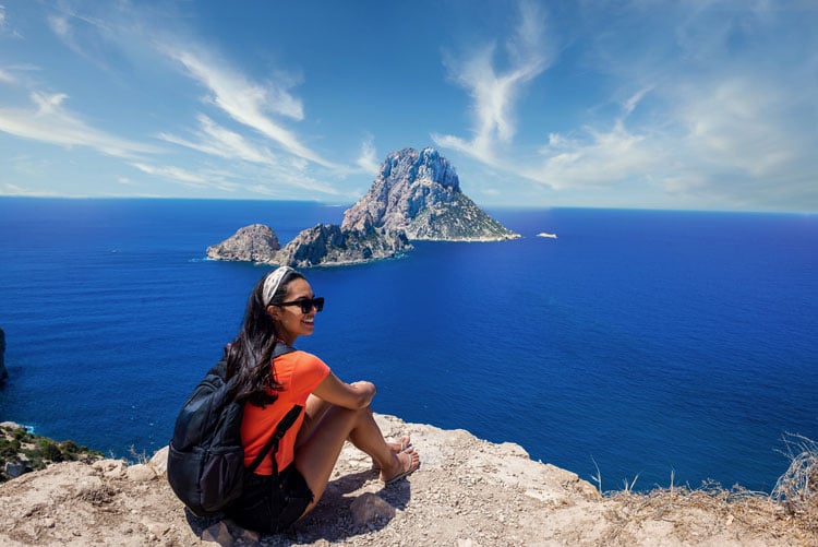 A woman in an orange shirt sits on a rock by the sea in Ibiza - one of the best places to hike in February