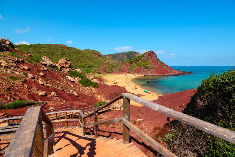 A wooden boardwalk along colorful cliffs by a beach in Menorca - one of the best places to hike in February