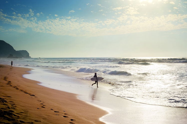 A person walking towards the sea with a surfboard in the Algarve - the best places to visit in May